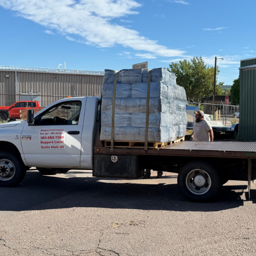 Flatbed delivery truck carries a large shrink-wrapped pallet outside an industrial building, with a worker nearby beneath a bright blue sky.