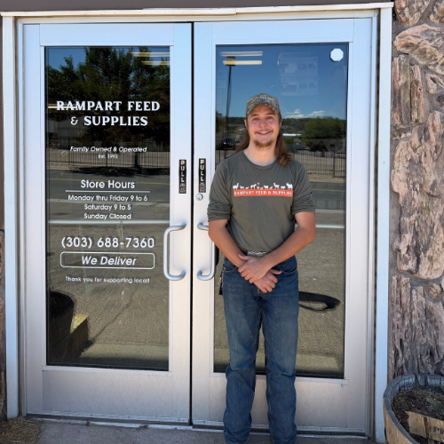 Smiling employee stands outside Rampart Feed & Supplies, a local pet and farm store, beside glass doors displaying hours, phone number, and delivery service.
