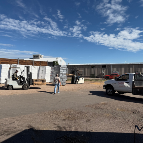 Industrial yard with workers operating forklifts near stacked pallets, a service truck, and warehouse buildings beneath a bright blue sky.