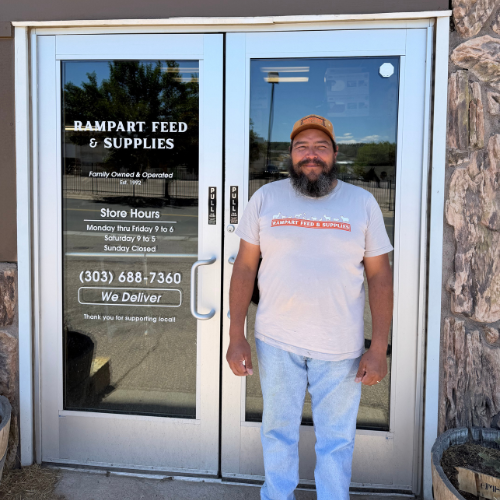 Smiling bearded man in a cap stands outside Rampart Feed & Supplies, beside glass doors showing store hours, phone number, and delivery notice.