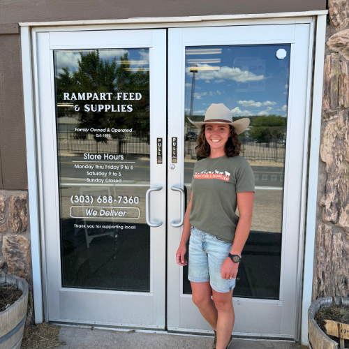Smiling woman in a cowboy hat stands outside Rampart Feed & Supplies, a family-owned store, beside glass doors listing hours and delivery.