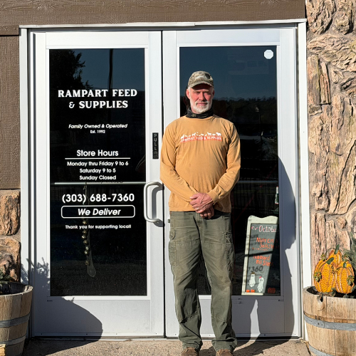 A man stands outside Rampart Feed & Supplies, beside glass doors listing store hours and delivery service; stone walls and planters give the shop a rustic, welcoming feel.