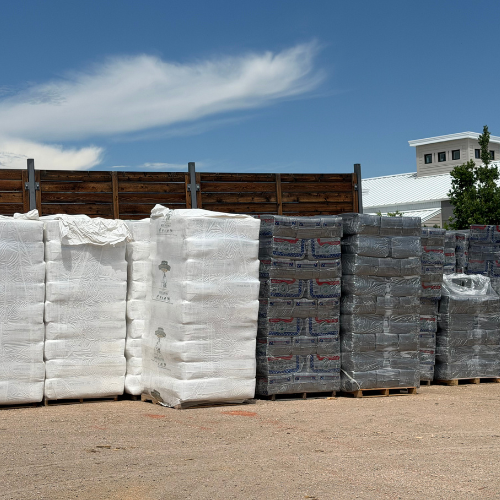 Outdoor building-supply yard with pallets of white wrapped bags and gray bricks stacked by a wooden fence under a sunny blue sky.